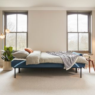 A blue sofa bed with a mattress topper and beige bedding on top in a room with two large windows