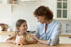 A woman talks to a young girl with a piggy bank sitting on a counter in front of them. 