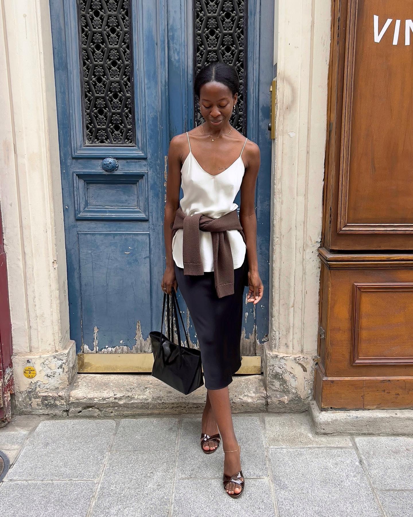 An image of @sylviemus_ wearing a navy slip skirt with a white silk camisole and brown cardigan while in Paris.