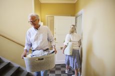 A mature man carries a laundry basket out of the laundry room while his wife is walking in. 
