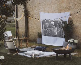 An outdoor setup with round black firepit, woven legless chair, picnic blanket and outdoor cushions, with festoon lighting decor above