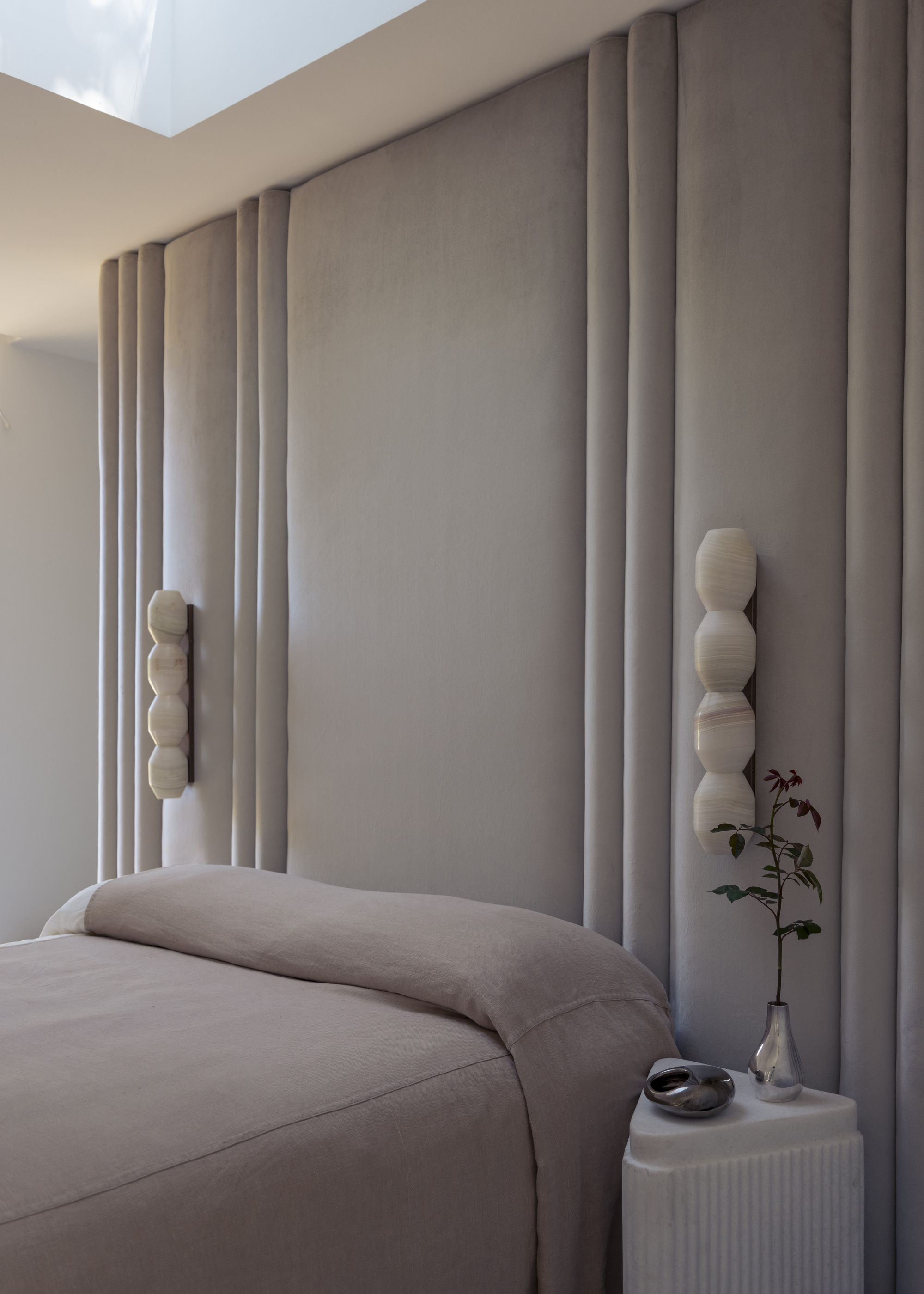 Image of a neutral bedroom with rose-pink undertones. The back wall is covered in fabric, and there are two sconces on the wall. The bedding is rose pink and there is a white marble side table.