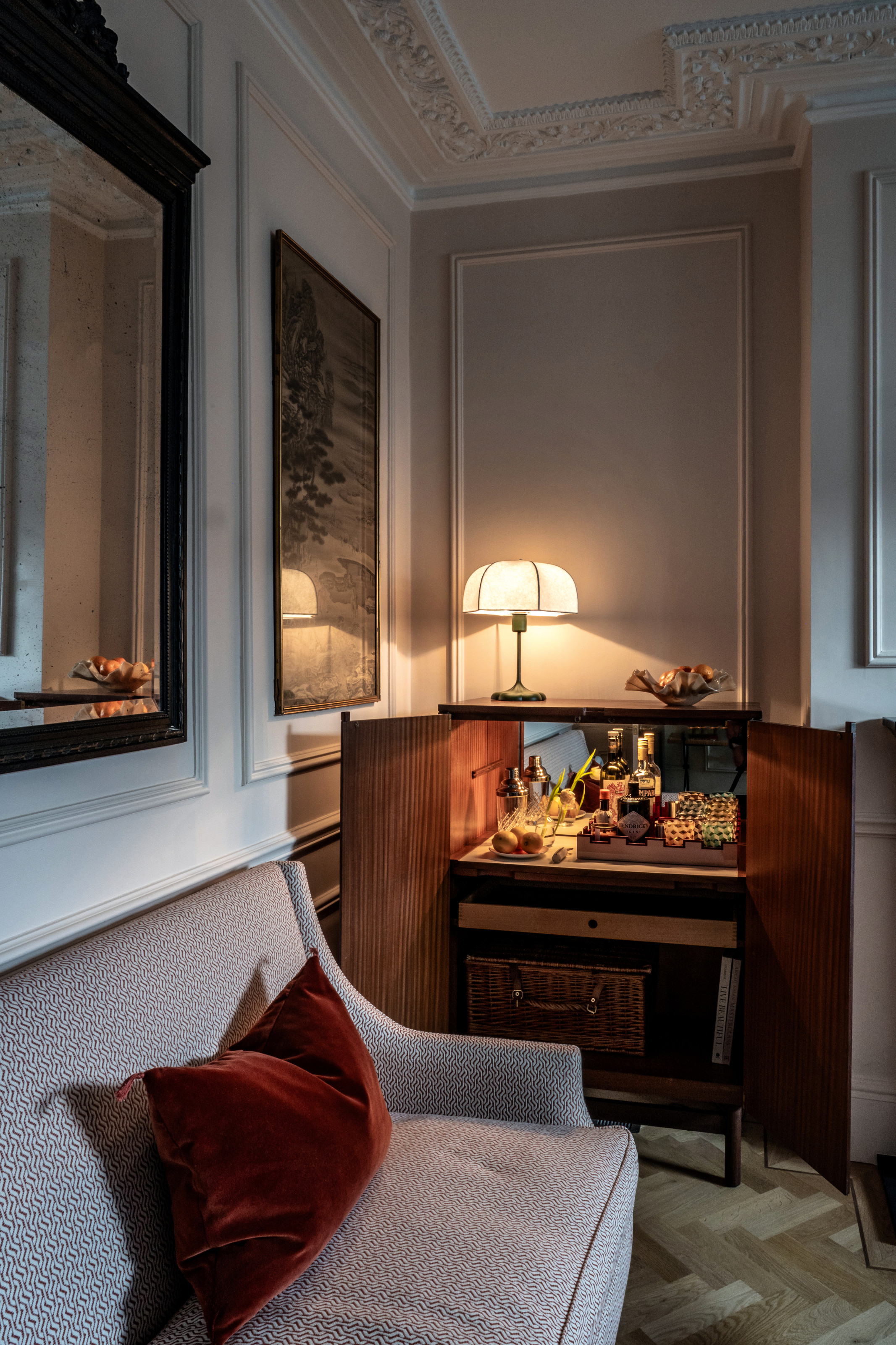 A corner of a room with a home bar in a wood cabinet, with a table lamp on top, next to a sofa with small-scale patterned upholstery