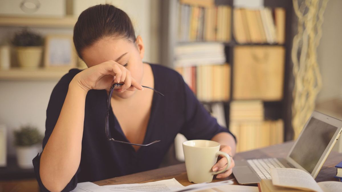 woman at a desk surrounded by papers and a laptop holds her head in one hand and a cup of coffee in the other