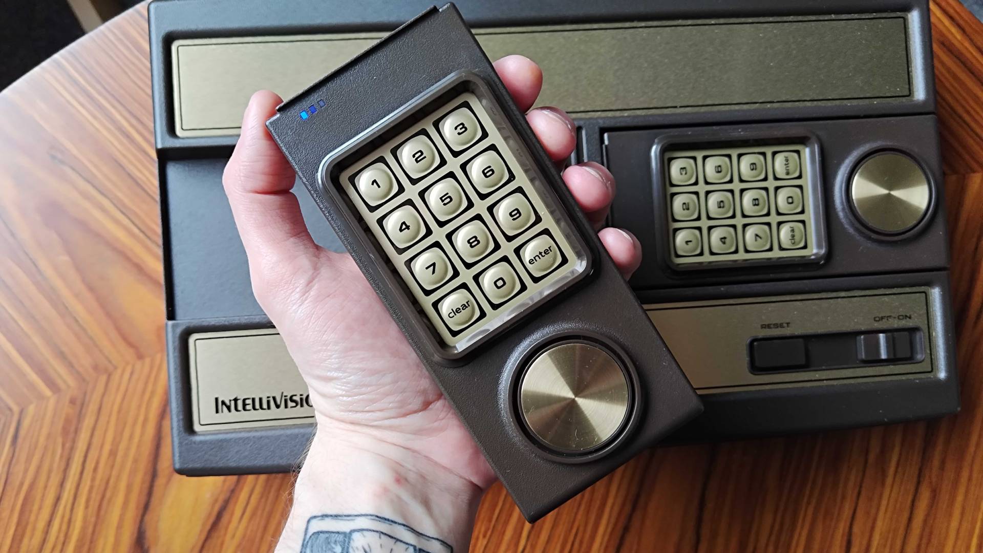 Hand holding Intellivision Sprint controller over console sitting on woodgrain table.