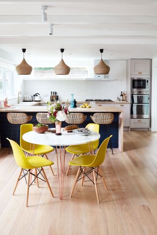 An open plan kitchen with a white round table, yellow chairs, a navy blue kitchen island, and rattan lights over these