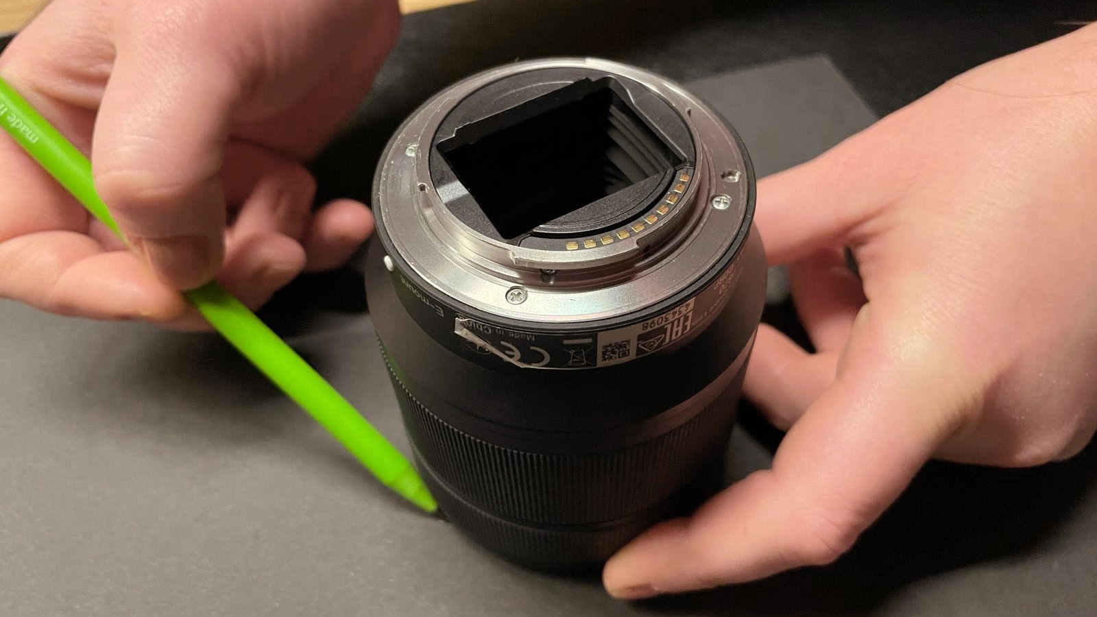A hands-on view of a camera lens with a green pencil beside it, focusing on the lens mount and aperture mechanics