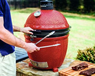 A man wearing a blue t-shirt and cream pants holding metal tongs and opening a red Kamado Joe barbecue smoker grill