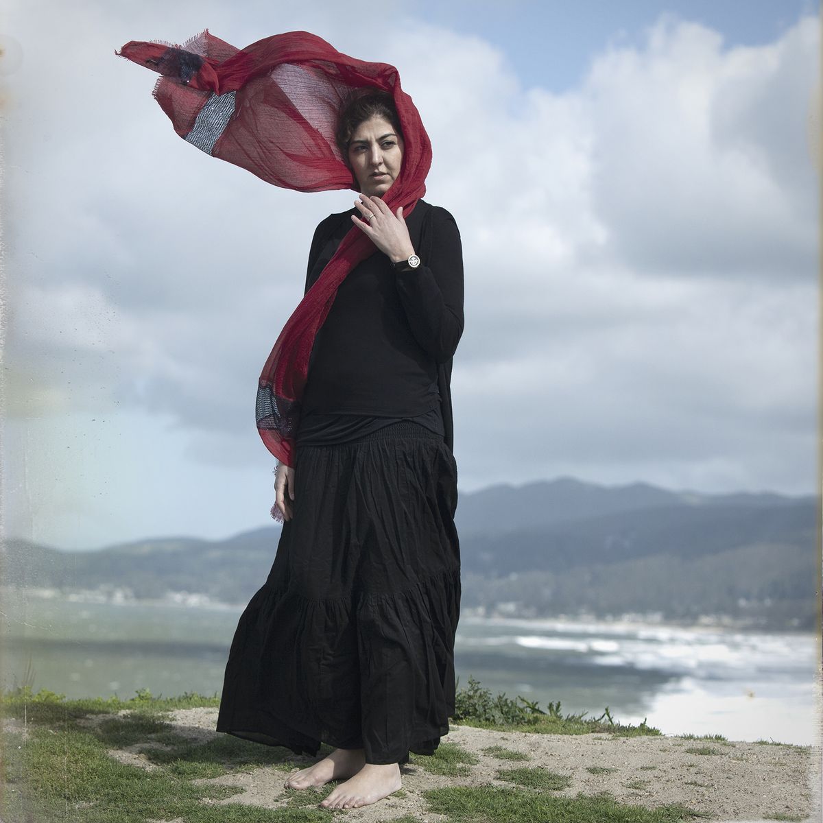 A woman stands barefoot on a grassy hill, wearing a black outfit and a flowing red scarf, with ocean waves and mountains in the background
