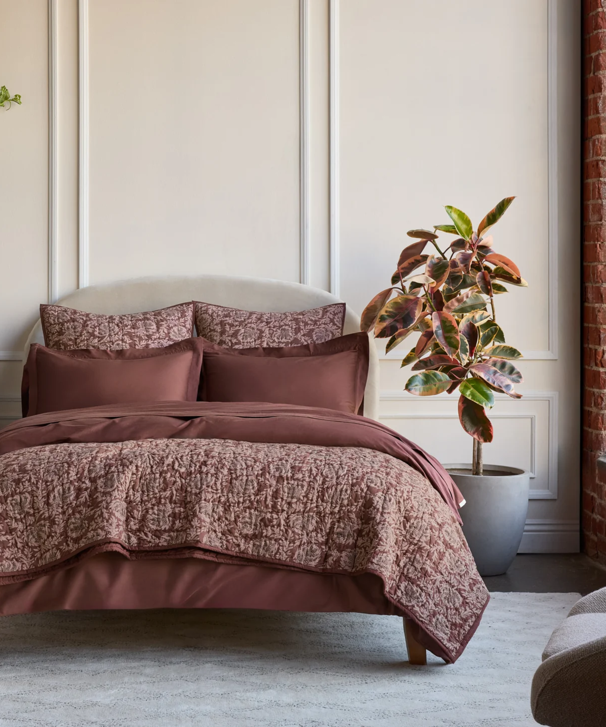 Moody, garnet Coyuchi bed sheets photographed in a bright, white bedroom alongside an oversized plant