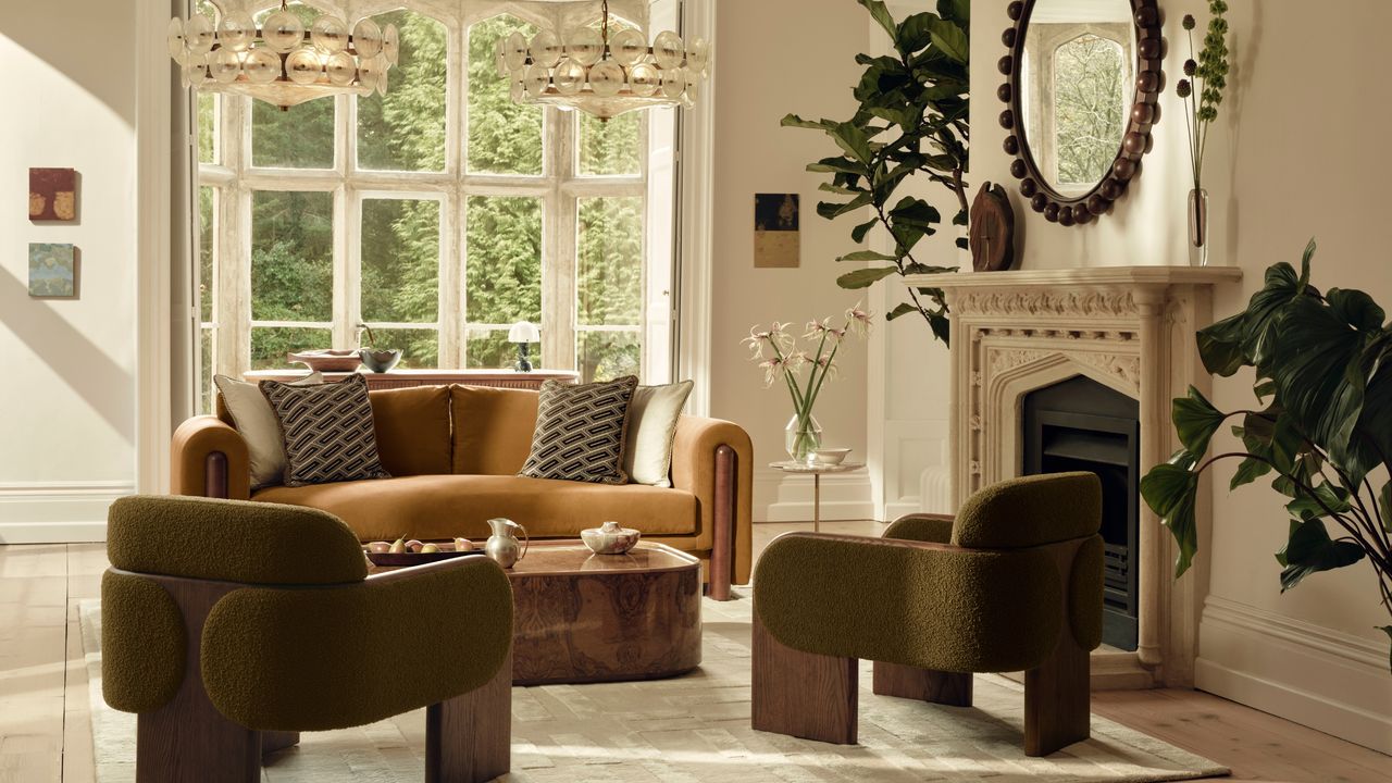 Image of a light, warm beige living room with two olive green accent chairs and a mustard orange sofa. There is a large marble fireplace with a bobbin mirror hanging over it to the side of the room.