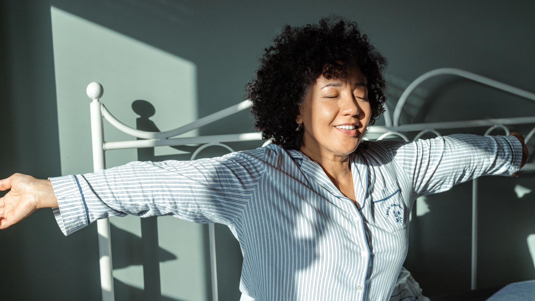 Woman in bed stretches her arms out to the sides, with sunlight hitting her face