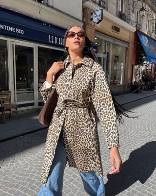 A influenciadora de moda francesa Lena Farouil posa em uma rua de Paris usando óculos escuros quadrados, um casaco com estampa de leopardo na cintura, bolsa de ombro marrom e jeans