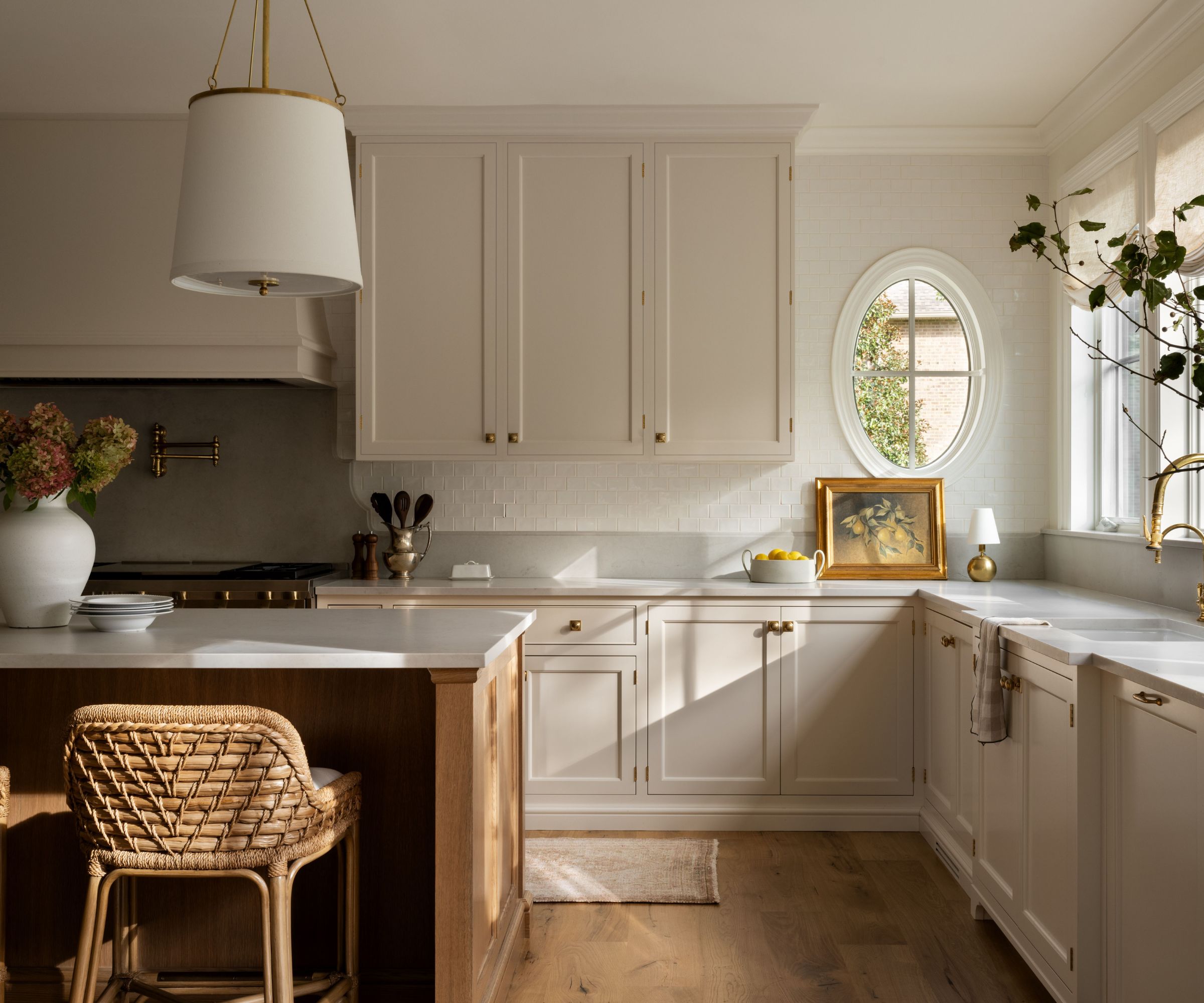 A neutral kitchen with an oval window, curved backsplash, rounded-back counter stools, and oversized pendant lights