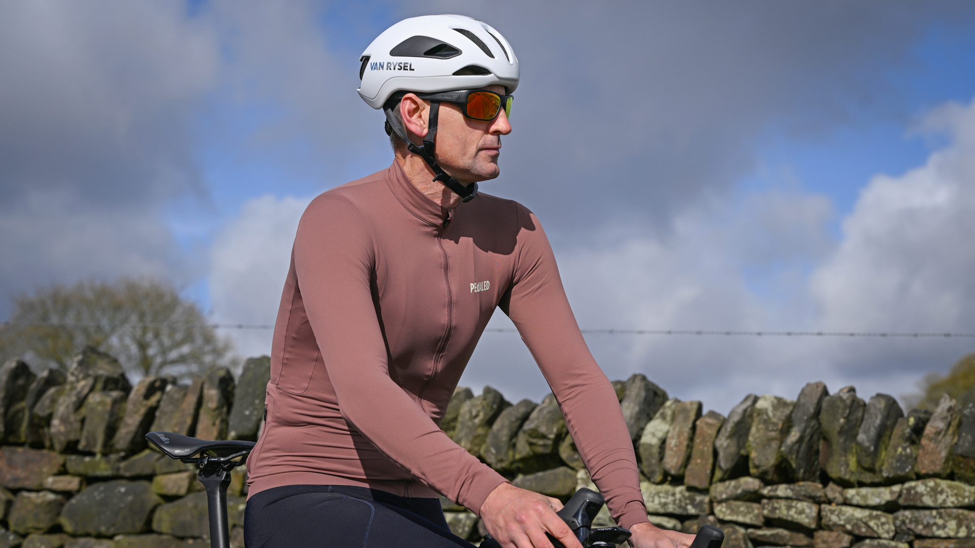 Man sitting on a bike wearing a white helmet, sunglasses and a long sleeved purple jersey in front of a drystone wall
