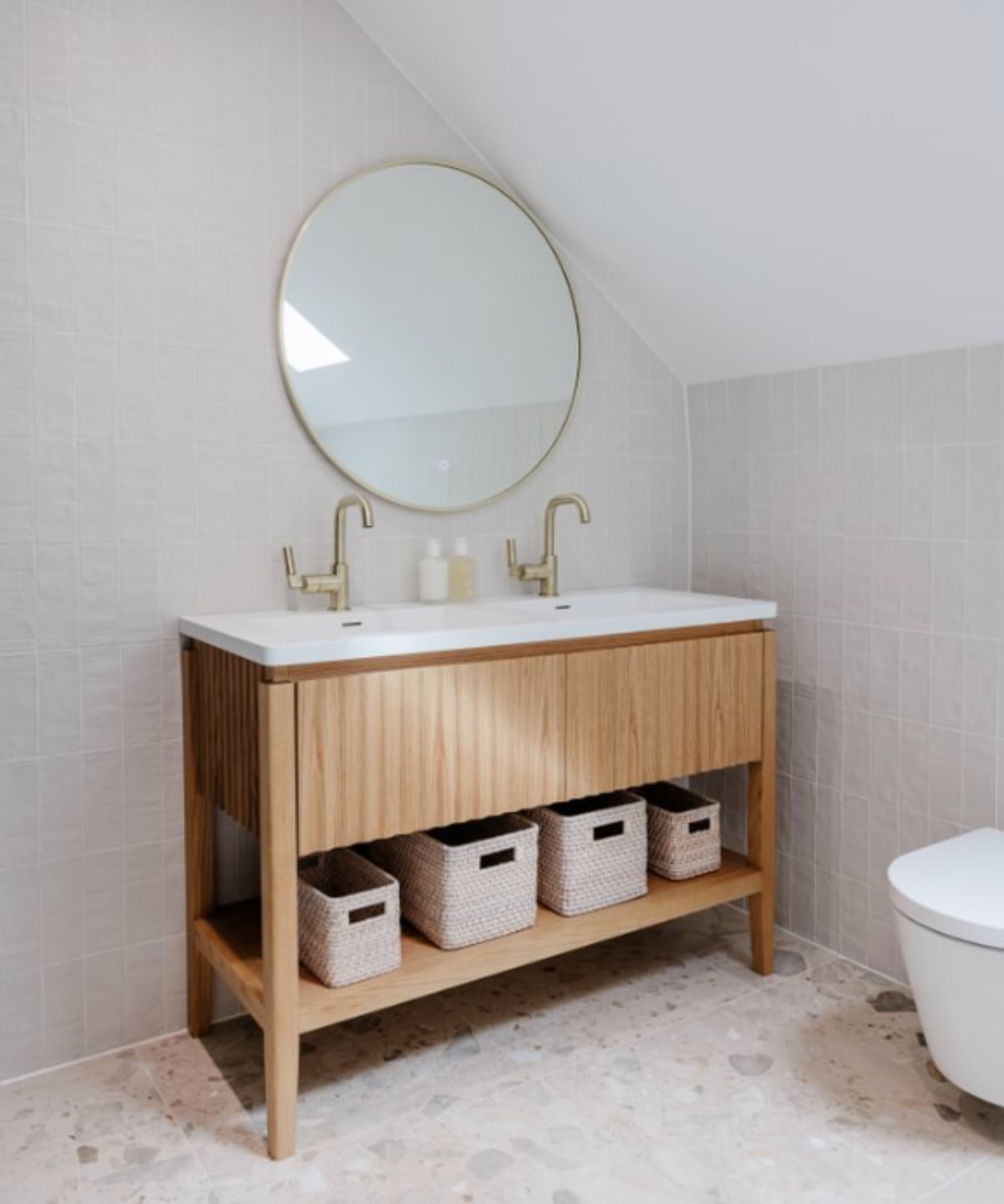 A light attic bathroom with soft grey square wall tiles and a terrazzo floor, featuring a freestanding oak vanity unit with a double white basin, two brass mixer taps and three wicker storage baskets on the lower shelf, beneath a large round brass-framed mirror.
