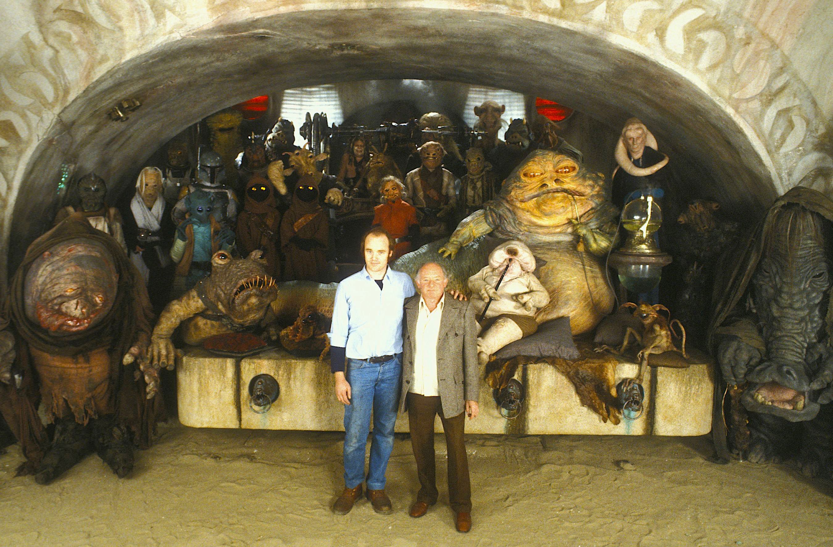 two men pose on a movie set surrounded by actors in alien costumes