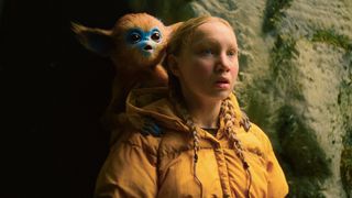 A girl wearing a yellow jacket looks shocked with a creature on her shoulder