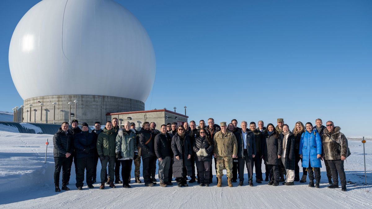 a group of military personnel stand on snowy ground under a blue sky, with a white spherical radar facility in the background