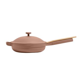 A neutral pink colored Our Place Always pan with a lid on top and a handle with a wooden spatula on it