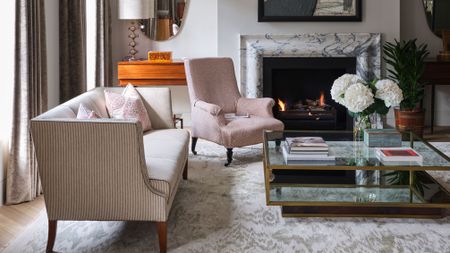 An elegant living room features a white marble fireplace with a crackling fire as its focal point, framed by a mix of classic and contemporary furniture. A light-striped settee with pink patterned pillows sits in the foreground, angled toward a brass and glass tiered coffee table holding stacks of art books and white hydrangeas. Beside the fireplace, a dusty-pink upholstered armchair adds a soft touch of color, while a warm wood console table with a decorative lamp and a large green potted plant complete the sophisticated, layered space.