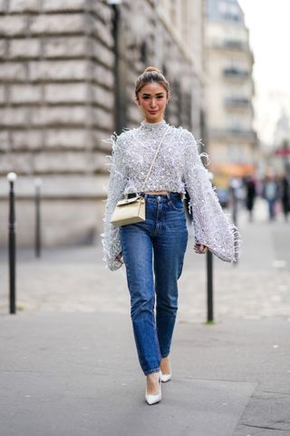 PARIS, FRANCE - MARCH 05: Heart Evangelista wears silver and rhinestones earrings, a gray embroidered crystal and rhinestones with feathers long puffy sleeves high neck pullover, a pale yellow shiny leather crocodile print pattern Kelly crossbody bag from Hermes, navy blue denim large pants, beige shiny leather crocodile print pattern pointed pumps heels shoes, outside Hermes , during Paris Fashion Week - Womenswear F/W 2022-2023, on March 05, 2022 in Paris, France. (Photo by Edward Berthelot/Getty Images)