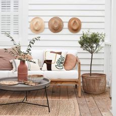 Sofa with cream and brown cushions, coffee table, olive tree in a basket on a wooden verandah deck outside a white wood clad house.