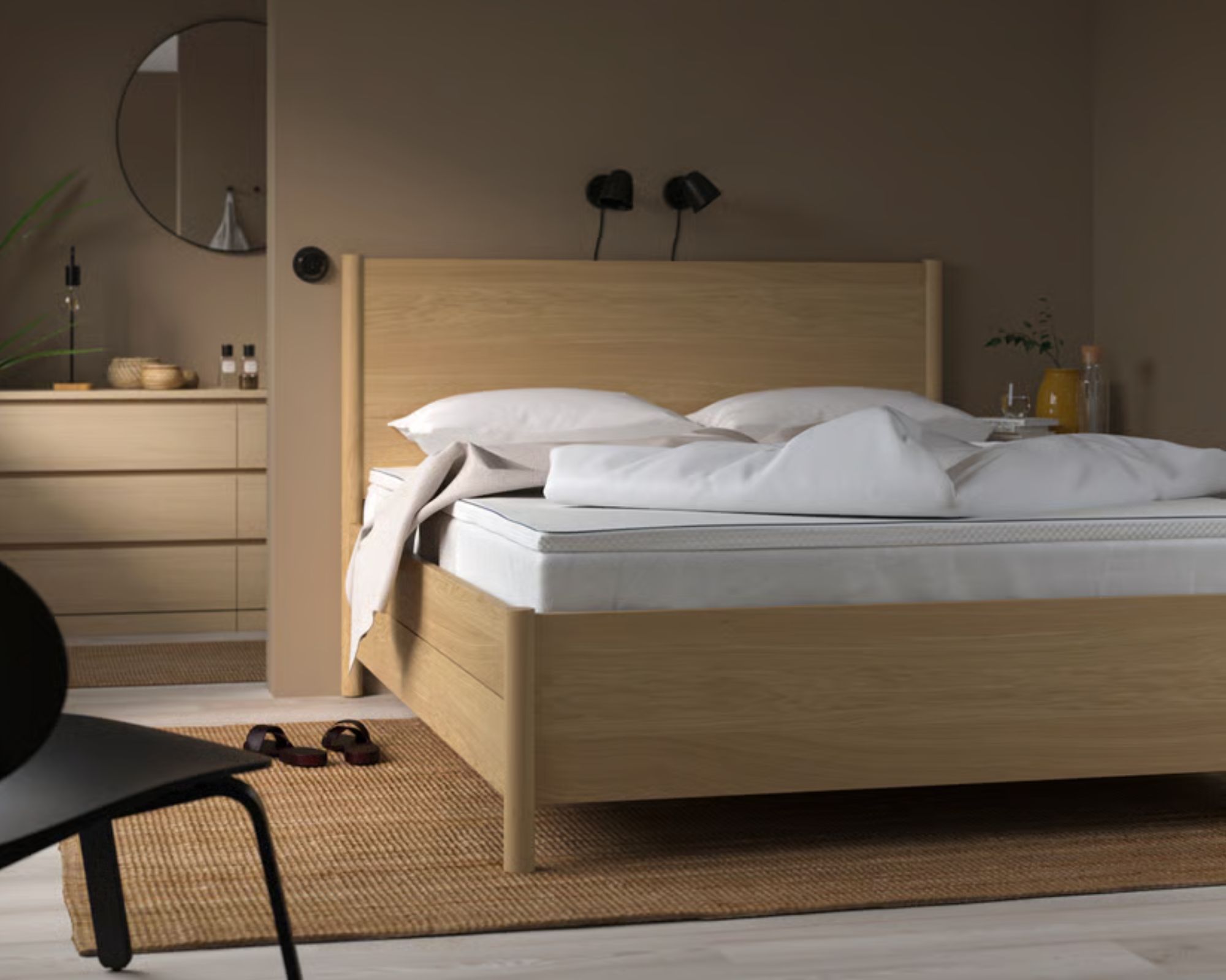 IKEA N&amp;Auml;STABERG Mattress Topper on a pine bedframe in a neutral brown bedroom with jute rug, black chair, and bathroom with drawers and circular mirror seen behind.