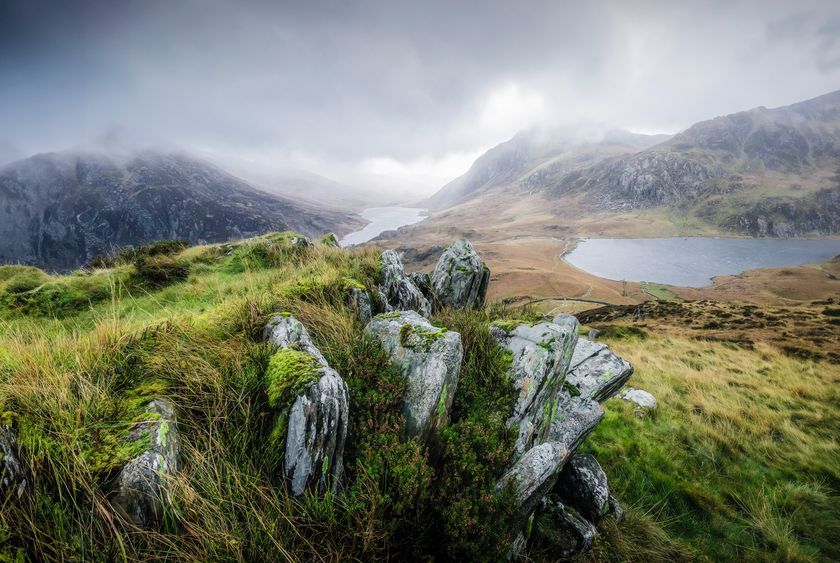 Big rocky foreground with mountains and lake in background, moody, rainy sky above