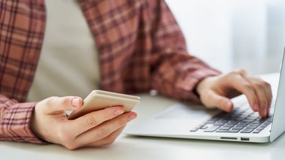 A man looks at his phone in one hand while his other hand is on the keyboard of his laptop.