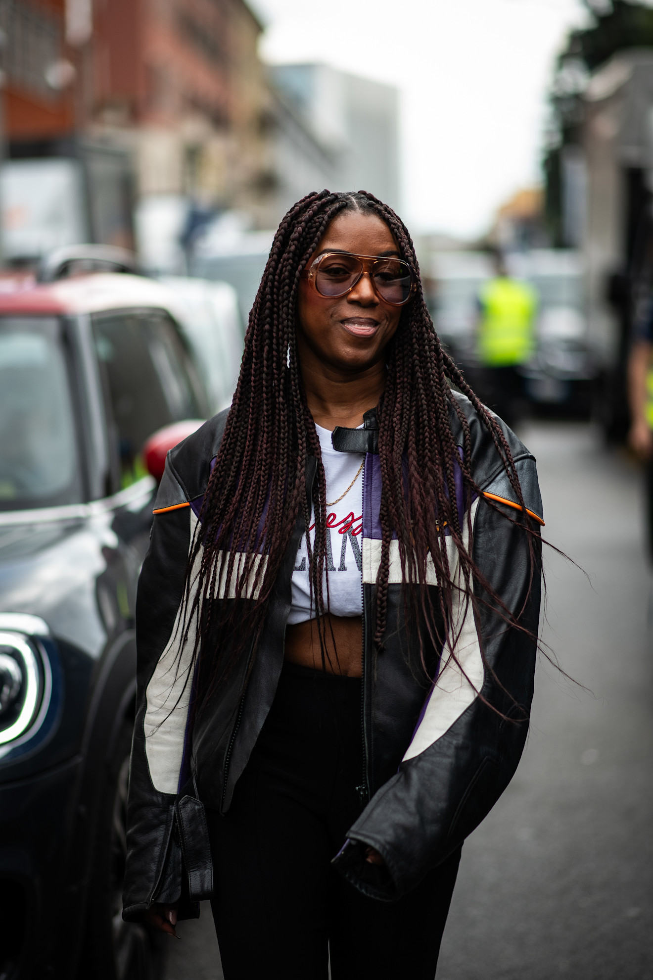 a woman with long box braids, a black and white bomber jacket and sunglasses facing the camera