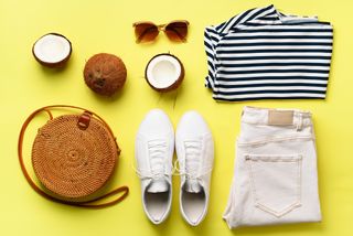 Female white sneakers, jeans, striped t-shirt, rattan bag, coconut and sunglasses on yellow background with copy space. Top view. Summer fashion, capsule wardrobe concept. Creative flat lay