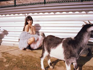 Singer Rebecca Black on a farm next to goats. She is crouched down with short bangs, and her brown hair is long and wavy. She is wearing a pink lace bra styled with a shaggy gray coat, a pink tutu, and leopard-print heels.