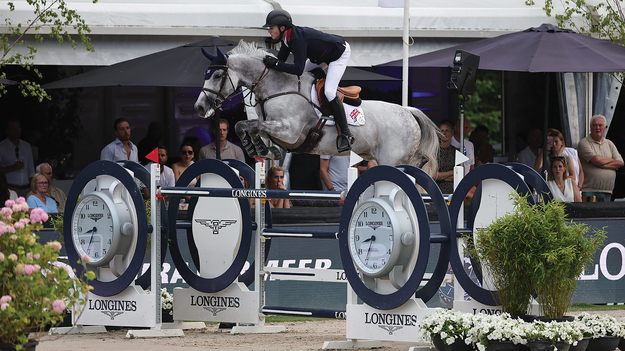 British showjumper Harry Charles rides a grey horse over a large navy and white Longines-branded oxer