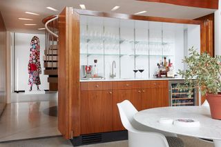 Wooden bar area with sink, glass shelves and a wine fridge next to a round table with white chairs
