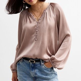 A model wears a pale pink voluminous blouse with jeans. She has lots of necklaces layered around her neck.