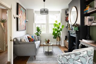Living room with wooden floors, white left-hand wall and green right-hand wall, black painted fireplace, round mirror, glass coffee table, grey sofa, pattern armchair and blinds in window
