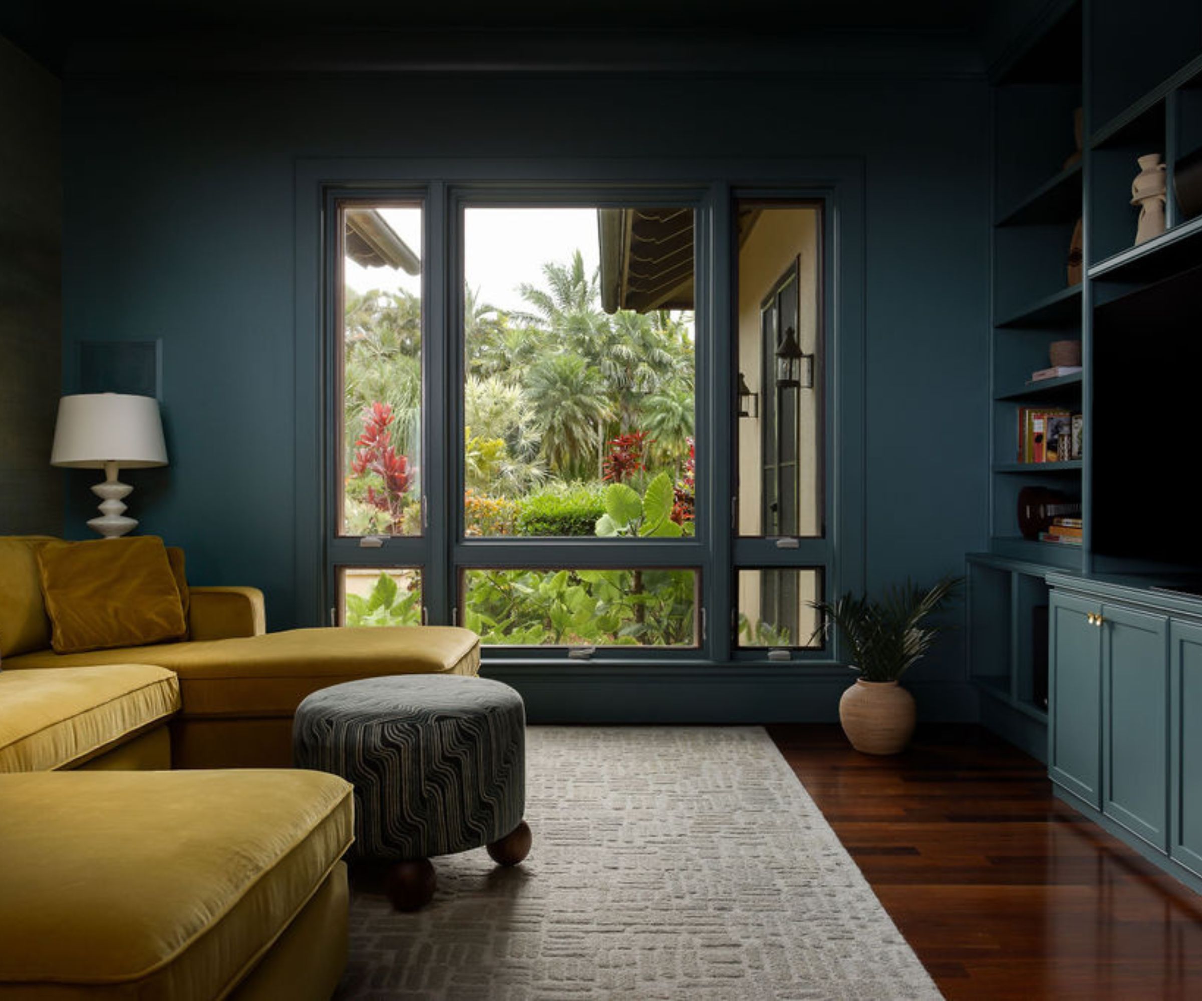 A modern TV room, color-drenched in dark teal with built-ins on one wall, dark wood flooring, a gray rug, and a large mustard-colored sectional sofa. A large window looking onto lush greenery outside