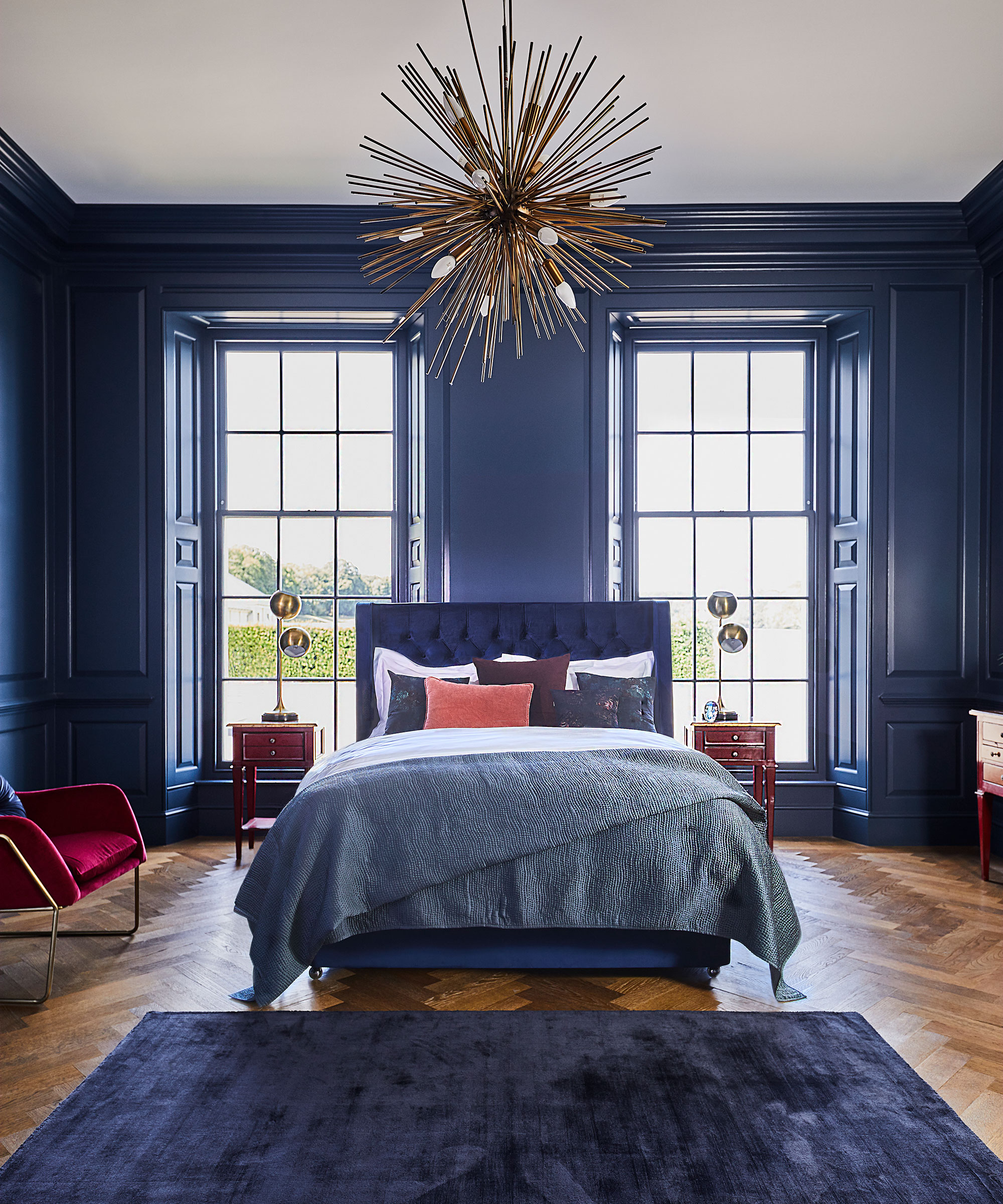 A blue bed in a large bedroom with dark blue walls and a statement pendant light