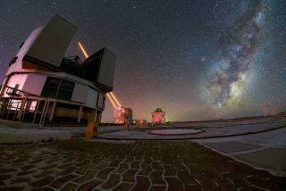 A foreground of curved bricks laid in a path leads to a round building with large section of roof opened. Behind it, another cylindrical building, shooting to brilliantly bright beams of light into the dark starry night sky. On the right, above the horizon, the arm of the Milky Way leans toward the top corner.