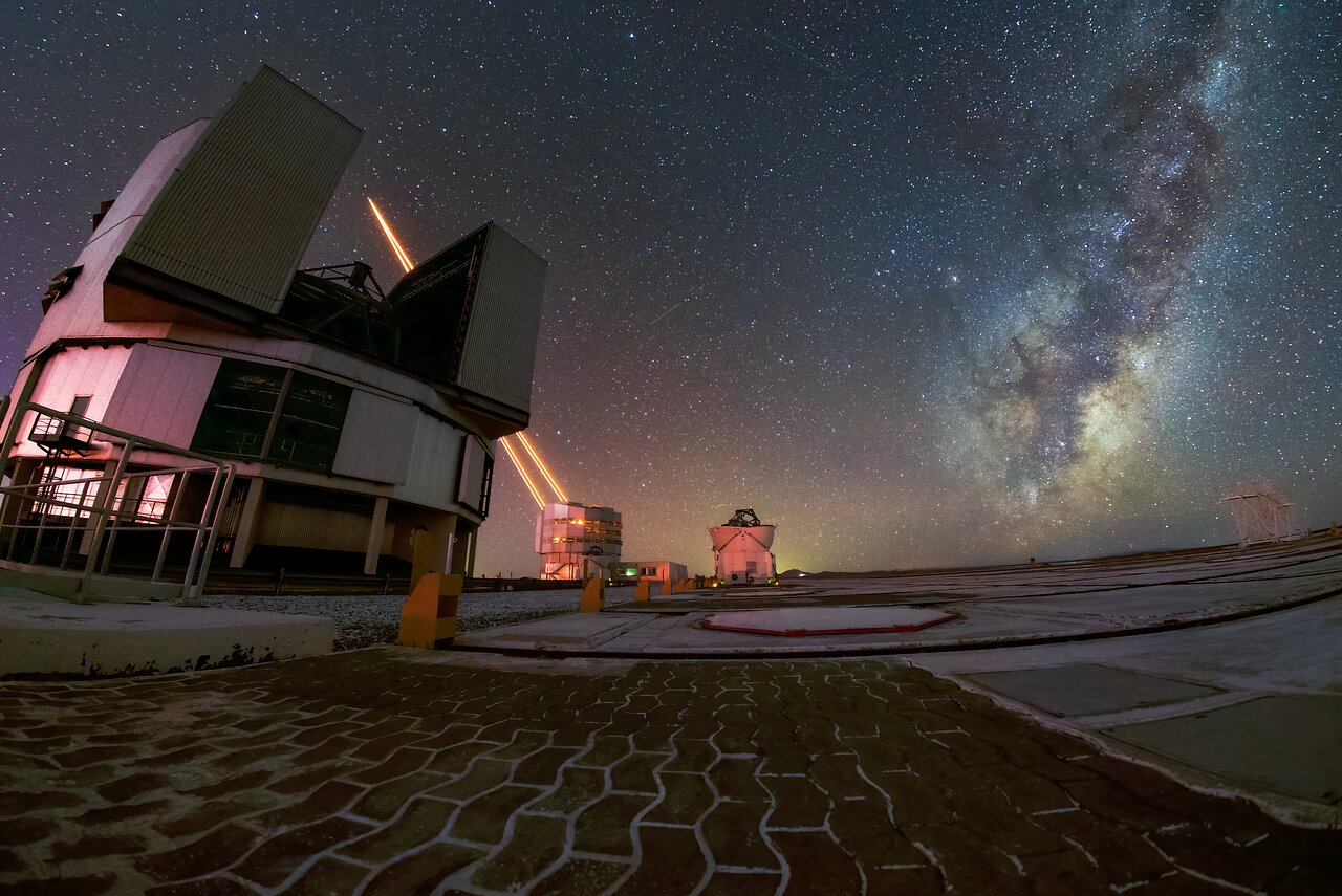 A foreground of curved bricks laid in a path leads to a round building with large section of roof opened. Behind it, another cylindrical building, shooting to brilliantly bright beams of light into the dark starry night sky. On the right, above the horizon, the arm of the Milky Way leans toward the top corner.