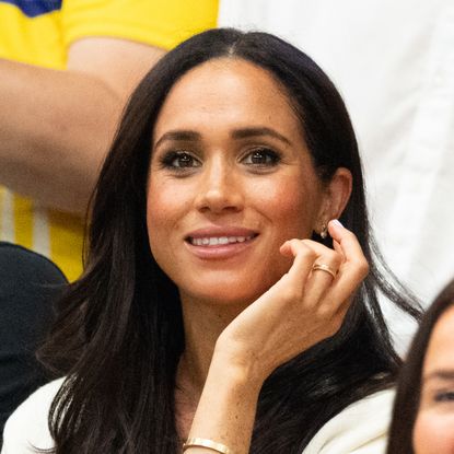 Meghan Markle wears a white jacket and smiles and has long black hair