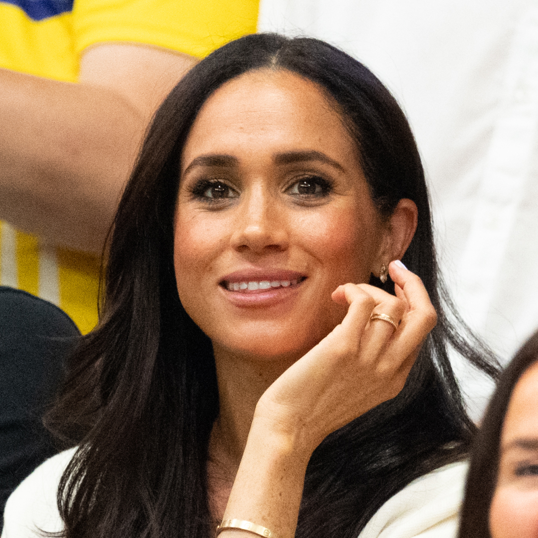 Meghan Markle wears a white jacket and smiles and has long black hair