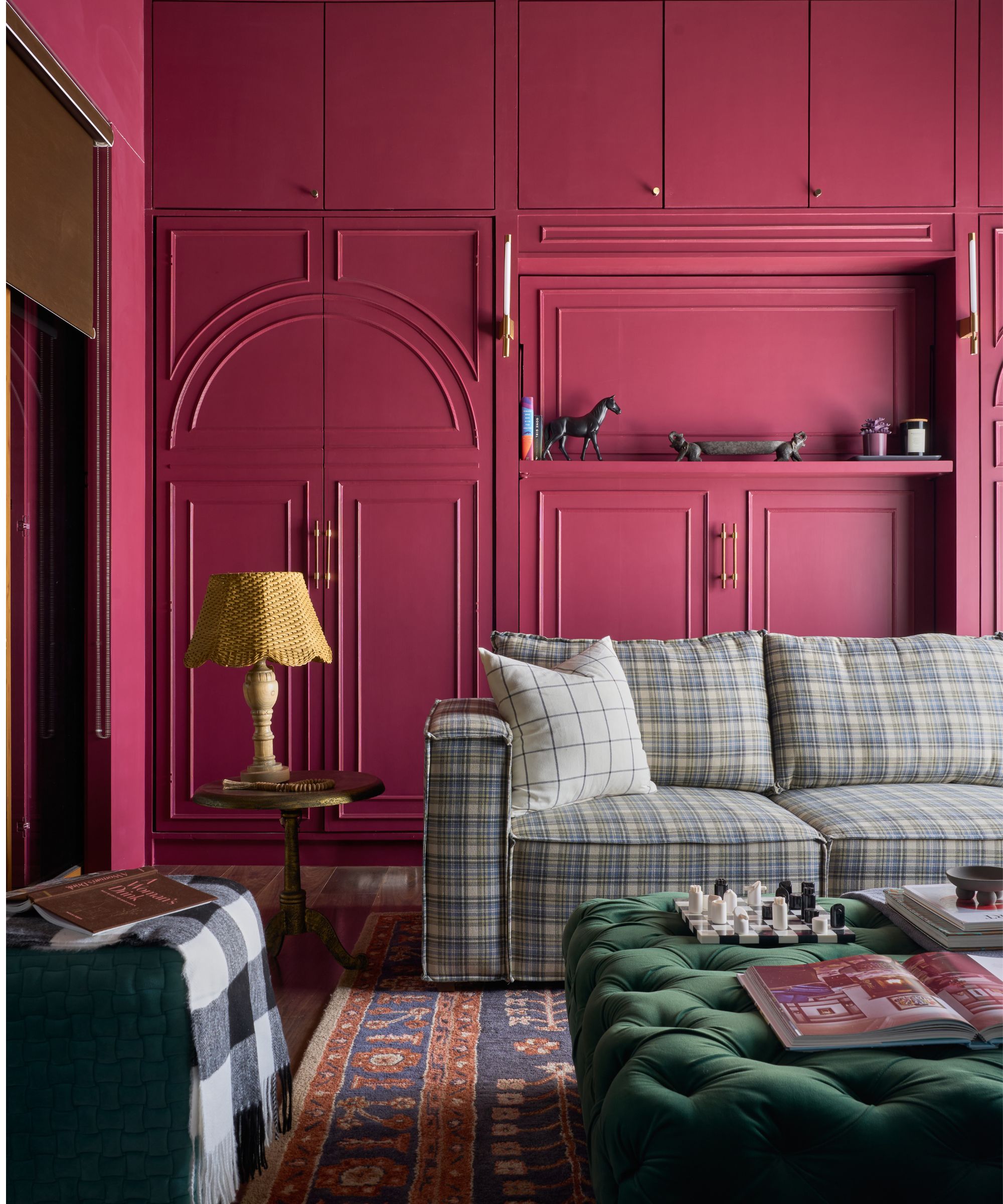 Living room with raspberry pink painted cabinetry and cupboards, a plaid blue sofa, a Persian rug, a green ottoman, and a gold side table with a yellow woven lamp on top