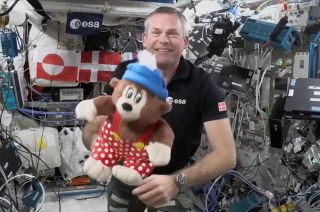 a man in a black polo shirt holds a plush bear inside the international space station.