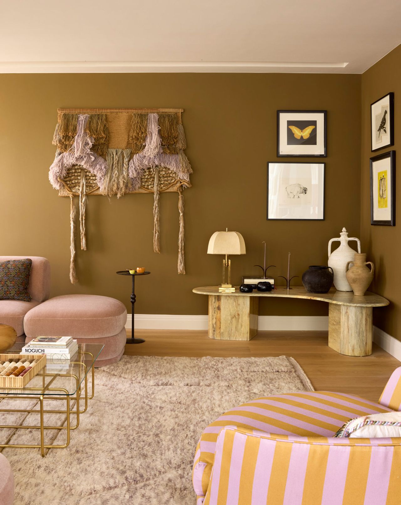 Image of a khaki colored living room with a more maximalist design style. There is a light pink sofa, a pink and orange chair, and a textured wall decor.