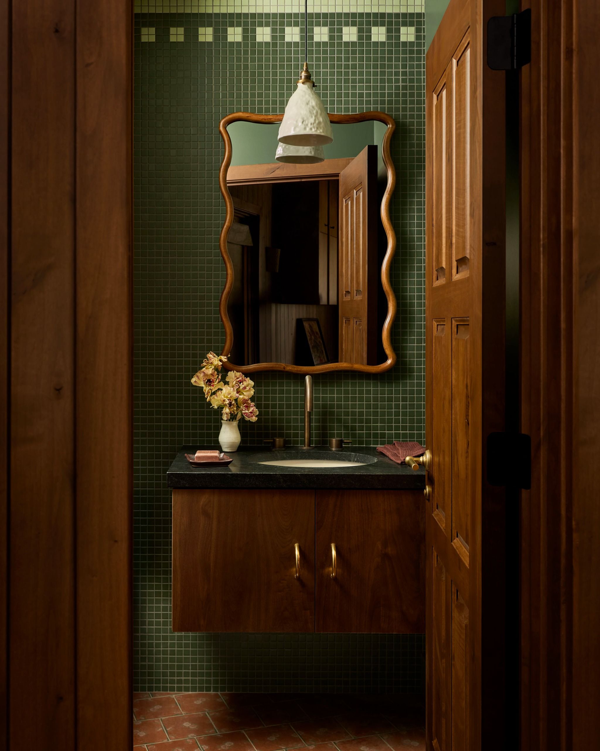 A powder room with green mosaic tiles on the walls, a floating wood vanity with a wiggle wood-framed mirror above it and a vase of flowers on the counter
