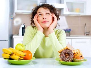 Young woman choosing between a bowl of fruit or a plate of unhealthy snacks.