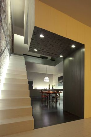 Cream stairs on the left with no banister leading up to another floor of the building. At the base of the stairs is a room with white walls and dark grey flooring. In the center of the room is a diing table with matching chairs