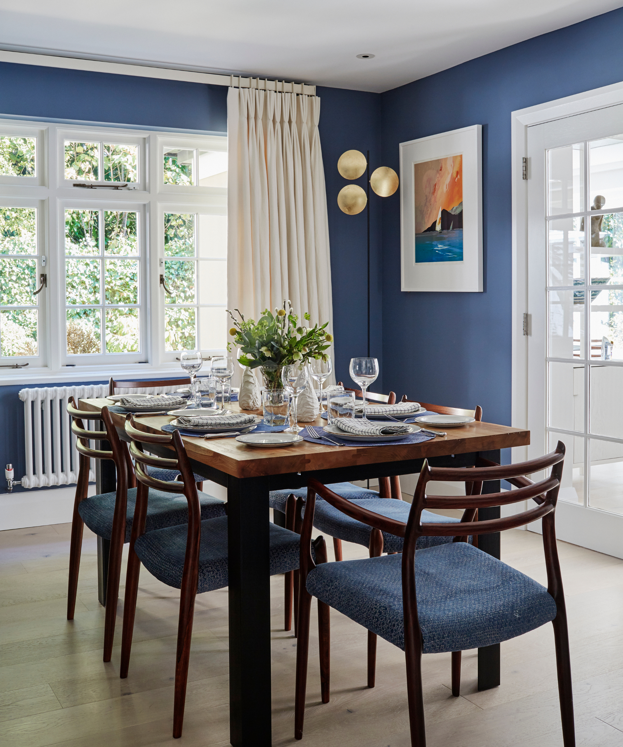 a blue dining room with dark wood furniture, a table set with six places, and a large set of windows with curtains in front and a radiator beneath